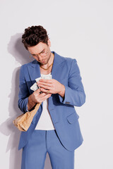 Stylish brunette man in blue suit counting money and holding paper bag on grey 