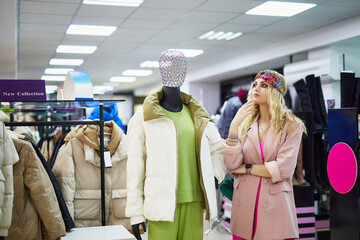 A young blonde chooses warm clothes in the store. A girl looks at women's clothes on mannequins. A...