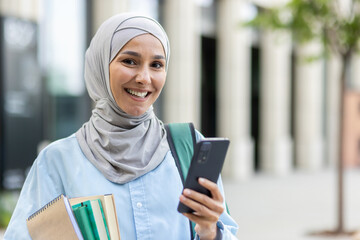 Smiling young Arab woman in hijab holding notebooks and a smartphone outside an office, symbolizing...