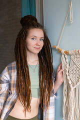 Young woman with dreadlocks weaves home decor from cotton thread macrame, looking at camera