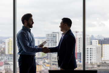 Fototapeta premium Two businessmen shake hands in an office, signifying agreement or collaboration, with a city skyline in the background.
