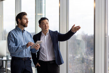 Two professional businessmen engaged in a discussion while one holds a tablet and the other points out of a window in a corporate office setting. - obrazy, fototapety, plakaty