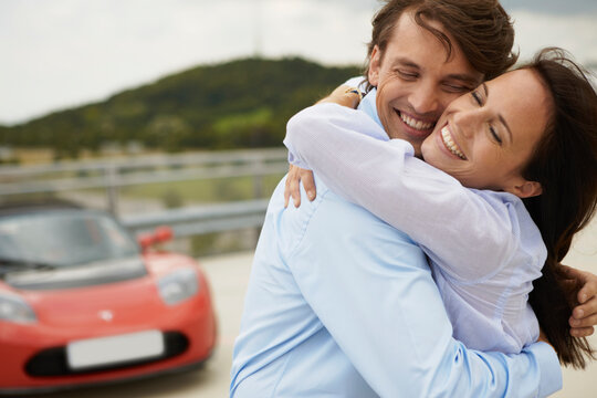 Couple in business attire hugging while on roof top car park with electric car in background. Munich, Bavaria, Germany