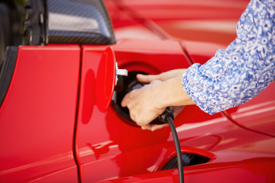 Woman plugging in her new red convertible electric sports car in front of modern house and garage. Munich, Bavaria, Germany
