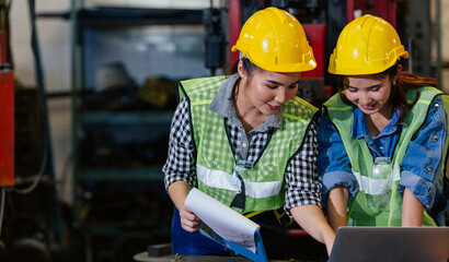 Two female engineers or technician worker wearing safety hard helmet discuss project in industry manufacturing factory. woman using laptop checking work, team colleague note on checklist document