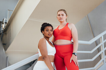 Portrait of two plus-size females in fitness attire looking at camera. Two plus-size athletes standing together outdoors relaxing after a workout.