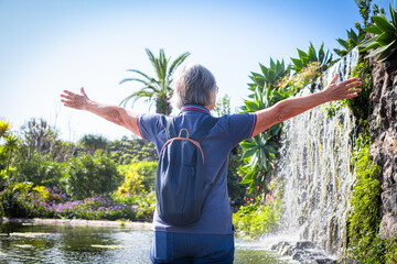 Rear view of caucasian mature senior woman with outstretched arms standing outdoors in a beautiful garden with tropical plants and waterfall