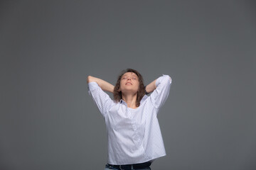 A young woman folds her hands behind her head and looks up smiling on a gray background.