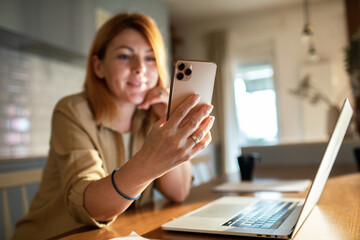 Obraz na płótnie Canvas Redheaded woman smiling and using smartphone in kitchen