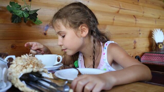 Little girl in white drinks tea by teaspoon in cafe 