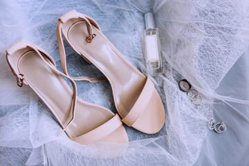 Wedding shoes for the bride. Elegant sandals of beige color for the bride stand on a white tulle. Wedding rings, earrings and perfumes. Bride's accessories. Wedding day