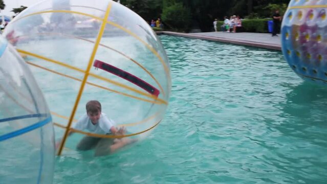 Boy teen flounders in large inflatable ball on water in pond