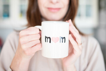 Caucasian woman, smiling happy about being a mother, holding a mother's day cup with "mom" written on it. Pink, airy background.