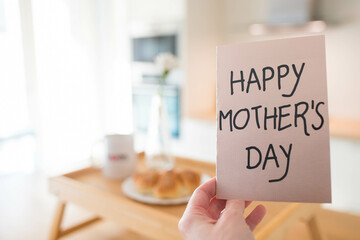 Mother's day still life with mom cup, a sweet pastry and a hand holding a greeting card, in airy indoor background.