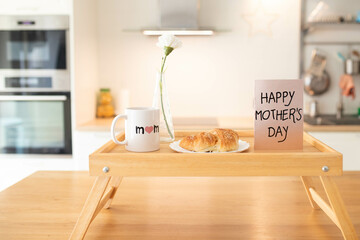 Mother's day still life with mom cup, a sweet pastry and a greeting card on a tray, in airy indoor background.