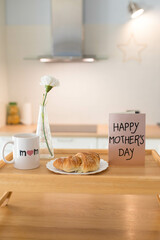 Mother's day still life with mom cup, a sweet pastry and a hand holding a greeting card, in airy indoor background. Vertical shot.