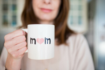 Caucasian woman, smiling happy about being a mother, holding a mother's day cup with "mom" written on it. Pink, airy background.