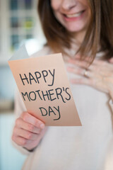 Caucasian woman, smiling happy about being a mom, holding a mother's day greeting card. Pink, airy background. Vertical shot.