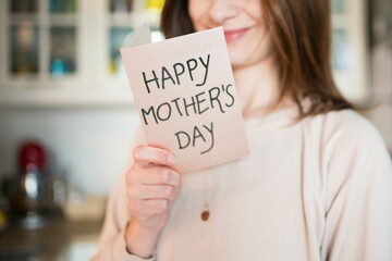 Caucasian woman, smiling happy about being a mom, holding a mother's day greeting card. Pink, airy background. 