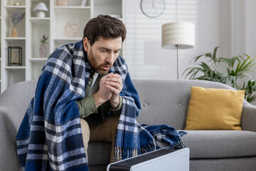A man appears cold and unwell, huddled under a checkered blanket on his couch at home, trying to...