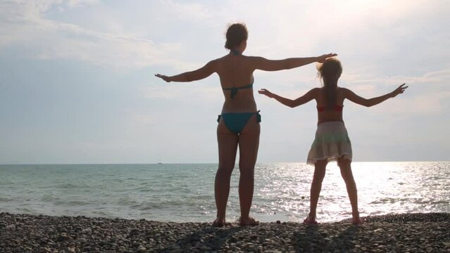 Mother and daughter stands arms to the side on the sea shore
