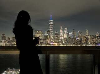 Fototapeta na wymiar woman looking at downtown manhattan nyc skyline (after sunset, night time) world trade center cityscape (high rise urban buildings) railing, hudson river, waterfront, bay, harbor new york city view