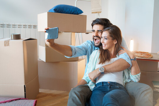 Joyous young excited couple making video call via phone with unpacked boxes during relocation. Happy young couple moving in new place and making a selfie with cardboard boxes.