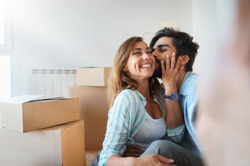 Loving young married couple hugging and kissing while taking selfie during relocation, surrounded with cardboard boxes around. - Powered by Adobe