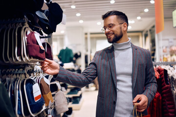 Young happy man buying knit hat in clothing store. - obrazy, fototapety, plakaty