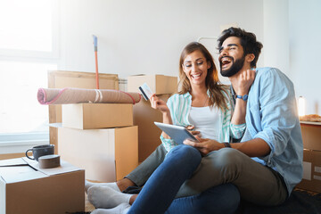 Enthusiastic millennial couple feeling excited using digital tablet, holding credit card, satisfied with online payment possibilities, rejoicing approved loan during relocation in new home. - obrazy, fototapety, plakaty