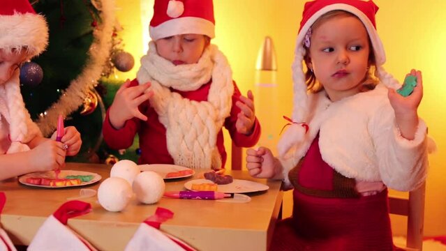 Two little girls and one boy in santa caps eat cookies at table