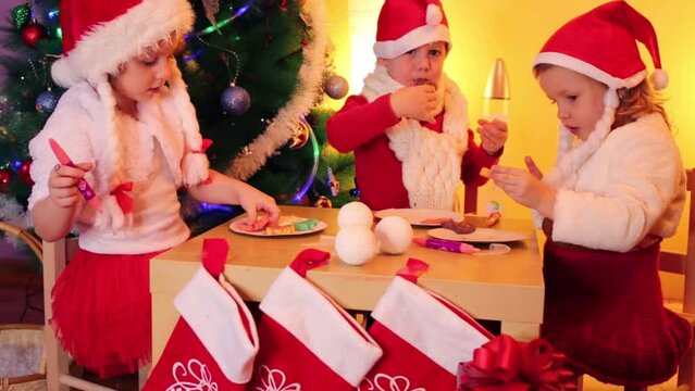 Two little happy girls and boy in santa caps eat colored cookies