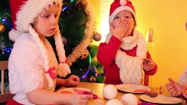 Two little happy girls and boy in santa caps eat cookies 