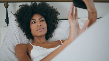 African woman posing for mobile selfie in bed. Smiling lady taking photo laying - obrazy, fototapety, plakaty