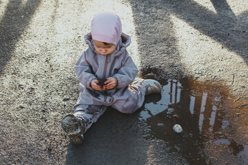 a child discovers the world, one small child in dirty clothes 1 year and 3 months old sits in a puddle and plays with a cone, the concept of childhood and walks - obrazy, fototapety, plakaty