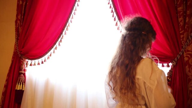 Beautiful girl in ethnic dress opens red curtains on window