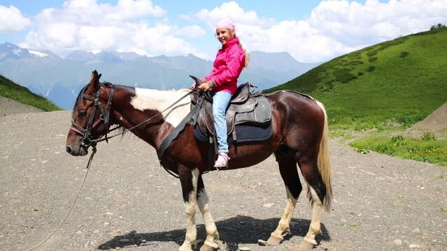 Pretty girl sits on horse among mountains at summer day