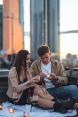 Fototapeta premium Romantic beautiful surprise on Saint Valentine's Day for a girlfriend. Loving young couple having dinner on a rooftop on sunset evening with candles, wine. Skyscrapers on background, urban cityscape