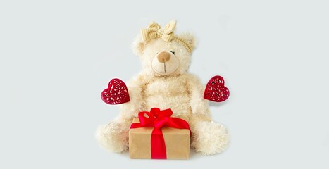 On a light background, a cute, soft teddy bear holds a gift and red hearts in its paws, a symbol of love.  Valentine's Day holiday concept.  Space for copying text.  Front view.