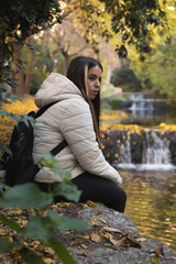 Un momento de paz y contemplación en un estanque otoñal con una cascada y árboles dorados, mientras una joven con chaqueta blanca disfruta de la naturaleza