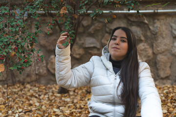 Una mujer joven y atractiva disfruta del otoño al aire libre, tocando con una mano una planta de muérdago que cuelga sobre su cabeza, mientras sonríe con gesto de felicidad y tranquilidad