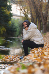 Mujer joven con chaqueta blanca expresa tristeza o relajación mientras se sienta junto a un lago rodeado de hojas de otoño