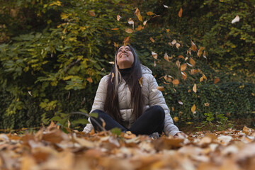 Mujer joven y hermosa con chaqueta blanca y pantalones negros se divierte lanzando hojas secas al aire en un parque otoñal rodeada de vegetación verde y colorida. - obrazy, fototapety, plakaty