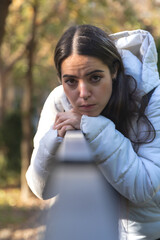 Mujer morena con chaqueta blanca y mochila negra se apoya en una barandilla en un parque soleado y mira al vacío con rostro borroso que refleja su estado de depresión