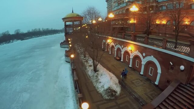 People walk by embankment near icy river and residential house