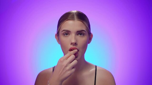 Kobieta na niebiesko-fioletowym tle patrzy centralnie w kamerę i wkłada do ust tabletkę, kt&oacute;rą następnie połyka. Ujęcie medyczne, spokojne i wyraziste, z naturalnym światłem i kolorem
