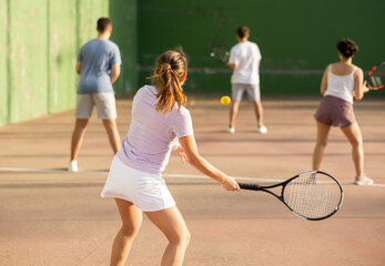 Rear view of young fit brown-haired woman playing frontenis team match on outdoor fronton court, swinging strung racket ready to hit small yellow rubber ball