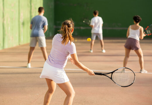 Rear view of young fit brown-haired woman playing frontenis team match on outdoor fronton court, swinging strung racket ready to hit small yellow rubber ball