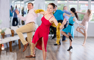 Happy brunette in stylish red dress decorated with golden tinsel dancing energetic upbeat jive paired with cheerful male partner during New Year party in dance studio