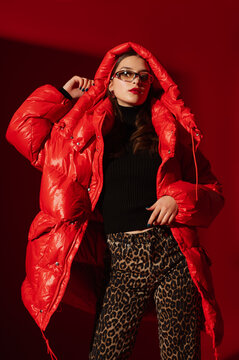 Fashionable confident woman wearing trendy oversized red puffer down jacket with hood, y2k sunglasses, leopard print jeans, black classic turtleneck, posing on red background. Studio fashion portrait